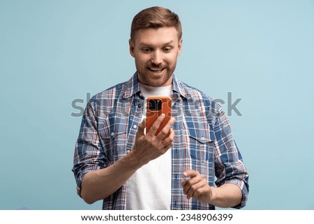 Man with smartphone smiles happily looks at screen isolated on blue wall. Male with phone smiling, expressing bright emotion of delight, jubilation, happiness, fun. Good news, surprise, joyful message