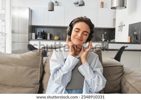 Portrait of beautiful, carefree girl listens to music in headphones with joyful smile, holds tablet, sits on sofa at home. Copy space