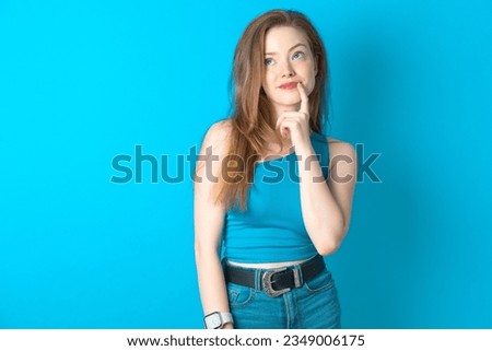 Lovely dreamy Young woman wearing blue T-shirt keeps finger near lips looks aside copy space. Royalty-Free Stock Photo #2349006175