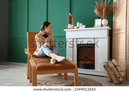 Young woman with mobile phone resting in armchair near fireplace at home