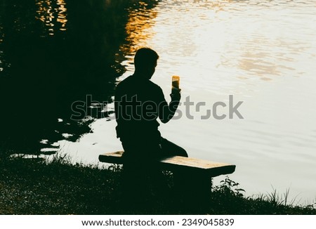 Portrait of a young man sitting on a bench and shooting video by the river Add color tones to make the picture look good with a lonely mood.