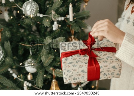 Woman hands opening stylish christmas gift with red ribbon close up at modern decorated vintage tree in scandinavian room. Merry Christmas and Happy holidays!
