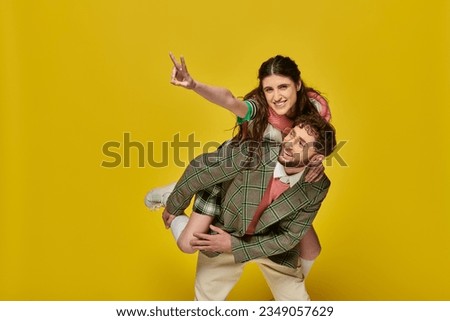 funny students, cheerful man piggybacking woman on yellow backdrop, v sign, college outfits, couple