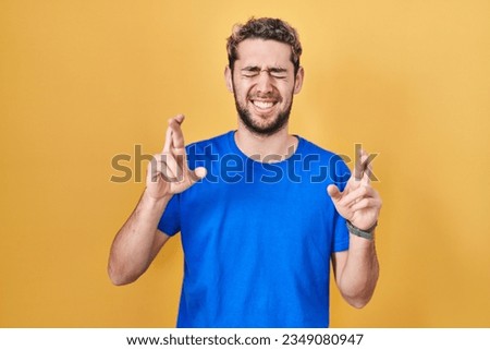 Hispanic man with beard standing over yellow background gesturing finger crossed smiling with hope and eyes closed. luck and superstitious concept. 