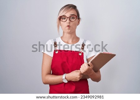Young blonde woman wearing waiter uniform holding clipboard making fish face with lips, crazy and comical gesture. funny expression.  Royalty-Free Stock Photo #2349092981