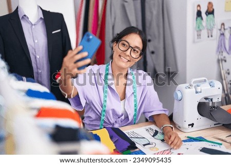 Young beautiful hispanic woman tailor using sewing machine make selfie by smartphone at tailor shop