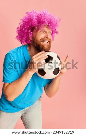 Emotional football fan, young caucasian man in funny wig watching soccer match and expressing emotions over pink background. Sport, fans, football