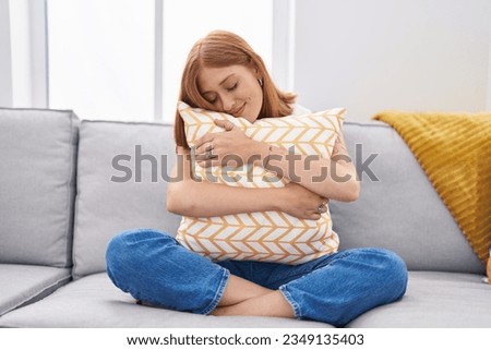 Young redhead woman hugging cushion sitting on sofa at home Royalty-Free Stock Photo #2349135403
