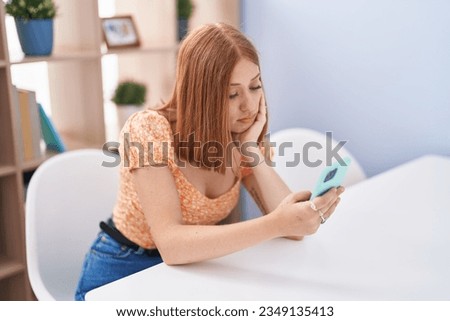 Young redhead woman using smartphone sitting on table at home