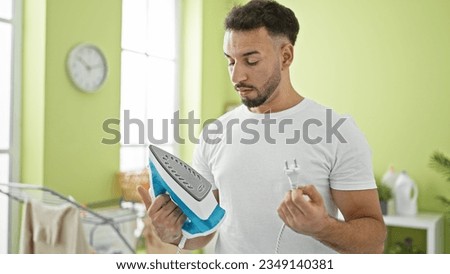 Young arab man holding ironing machine looking upset at laundry room