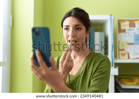 Middle age hispanic woman business worker using smartphone as a mirror make up lips at office