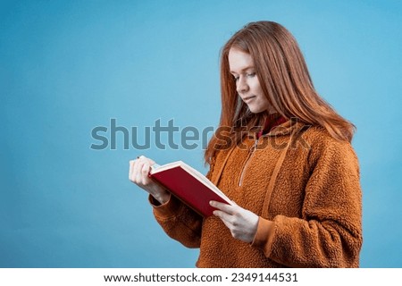 a young woman attentively reads a book standing on a blue background in the studio. reading books, money books and knowledge day Royalty-Free Stock Photo #2349144531