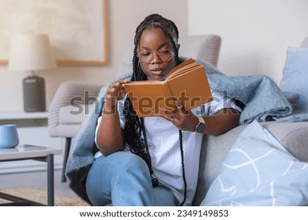 Close-up pensive dark-skinned girl reading a book, studying, sitting on the floor near the sofa, enjoying the home peaceful atmosphere in the morning. The concept of people and leisure, education.