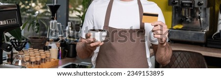 cropped view of barista in apron holding credit card and cup while working in coffee shop, banner