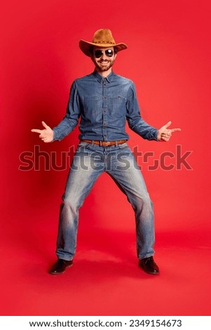 Excited young man in cowboy style outfit having fun, posing isolated over red background. Model in cowboy hat and denim clothes. Fashion, emotions concept. Looks happy, delighted Royalty-Free Stock Photo #2349154673