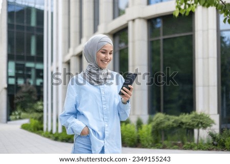 A young beautiful business woman in a hijab walks through the city, a Muslim woman holds a phone in her hands, uses an application on a smartphone, smiles, browses the Internet. Royalty-Free Stock Photo #2349155243