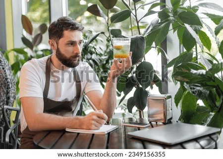 barista in apron holding lemonade and writing on notebook near laptop on table in coffee shop