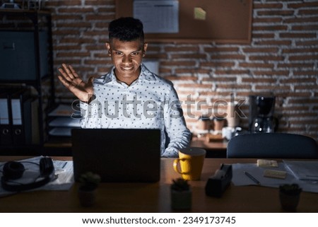 Young hispanic man working at the office at night smiling cheerful presenting and pointing with palm of hand looking at the camera. 