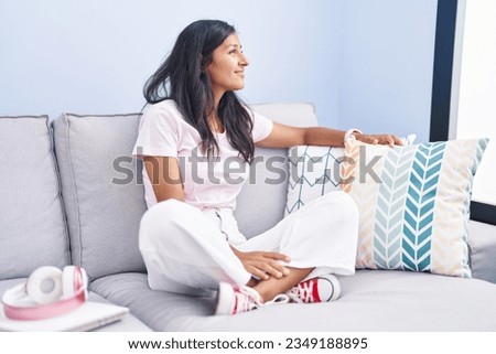 Young hispanic woman sitting on the sofa at home looking to side, relax profile pose with natural face with confident smile. 