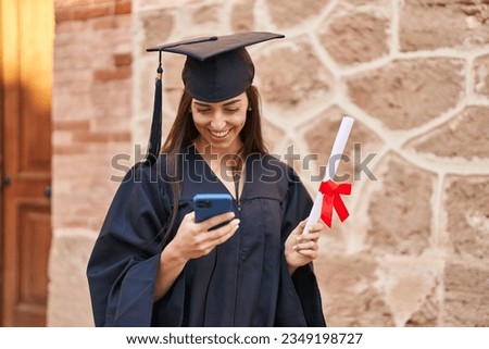 Young hispanic woman wearing graduated uniform holding diploma using smartphone at university Royalty-Free Stock Photo #2349198727