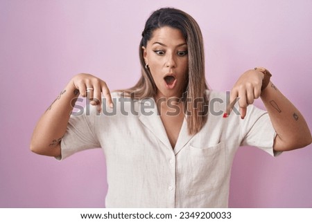 Blonde woman standing over pink background pointing down with fingers showing advertisement, surprised face and open mouth 