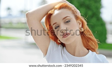 Young redhead woman standing with relaxed expression combing hair with hand at street
