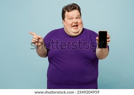 Young surprised amazed chubby overweight man wear purple t-shirt casual clothes hold use mobile cell phone with blank screen area point aside isolated on plain blue cyan background. Lifestyle concept