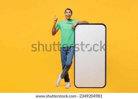 Full body smiling happy smart young man he wears casual clothes green t-shirt hat big huge blank screen mobile cell phone smartphone with area point index finger up isolated on plain yellow background