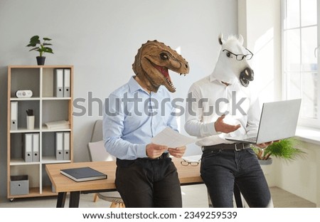 Funny animal people use computer at work. Team of 2 men in unusual masquerade dinosaur and horse masks standing by desk in office workplace, preparing creative business presentation on laptop device Royalty-Free Stock Photo #2349259539