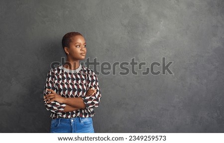 Portrait of attractive young African American woman who is thoughtfully smiling while standing against gray wall. Confident stylish casual woman looking towards free space for text. Banner background.