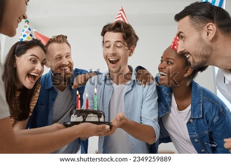 Portrait of happy laughing young people dressed in casual clothes and funny colored cone party hats celebrating birthday. Young beautiful birthday man is handed small cake with candles.