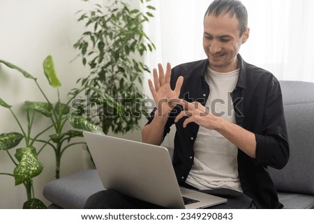 Deaf or hard hearing happy smiling young caucasian man uses sign language while video call using laptop while sitting on the couch at home.