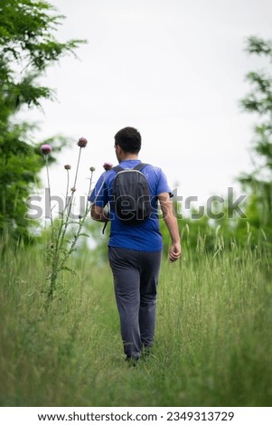 Professional portrait photo of a young man with backpack walking in a forest, tall grass around him, picture from behind 