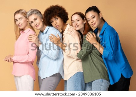 Group of beautiful smiling multiracial women wearing stylish colorful shirts looking at camera isolated on beige background. Portrait happy different ages fashion models posing for pictures in studio