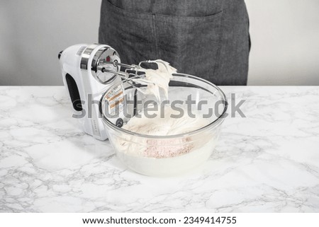 Whisking buttercream frosting in a glass mixing bowl with an electric hand mixer to prepare the peppermint buttercream frosting.