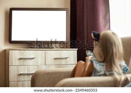 Back view of little girl kid 5-6 year old watching tv sitting on armchair with remote control, empty isolated screen. Cute girl child look movie on tv. Television addiction concept. Copy ad text space