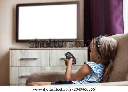 Rear view of little girl child 5-6 year old sitting on chair with remote control watching tv, empty isolated screen. Cute girl kid look cartoon on tv. Television addiction concept. Copy ad text space