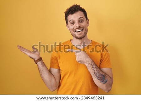 Young hispanic man standing over yellow background amazed and smiling to the camera while presenting with hand and pointing with finger. 