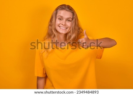 Young positive Caucasian woman blonde shows thumbs up and smiles broadly demonstrating full agreement with your opinion or satisfaction with quality of services rendered stands in orange studio.
