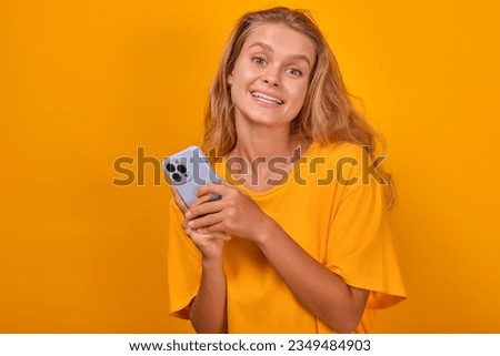 Young satisfied casual Caucasian woman with mobile phone in hand looks at camera and uses smartphone to exchange messages with friends or communicate in instant messengers stands in orange studio. Royalty-Free Stock Photo #2349484903