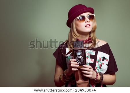 Studio portrait of teenage hipster girl wearing trendy hat and sunglasses with retro photo camera over olive copy space background