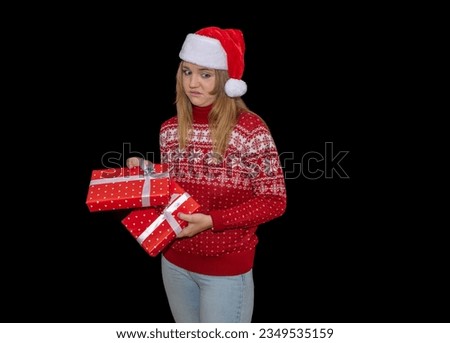 light-eyed blonde young girl looks at her christmas gifts with a pouting face. Blue-eyed woman holds a gift, dressed in a Santa Claus hat and a red Christmas sweater on a black background.