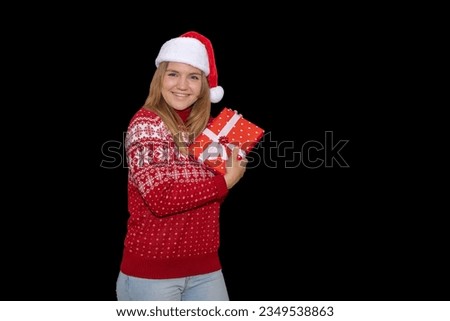 Young blue-eyed blonde girl with happy attitude holding some gifts on black background. Light-eyed woman in a Christmas sweater and Santa Claus hat looks at Christmas gifts with a face of illusion.