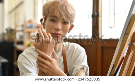 Portrait image of an attractive Young Asian teenage LGBT artist boy with colored hair showing his hands with a paintbrush, Doing artwork, Painting color on a canvas in the studio workshop.