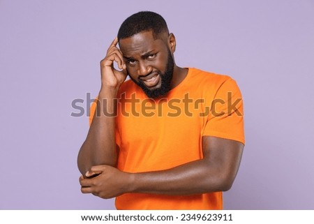 Preoccupied worried young man of African American ethnicity wearing basic empty blank orange t-shirt stand put hand on head looking camera isolated on pastel violet colour background studio portrait