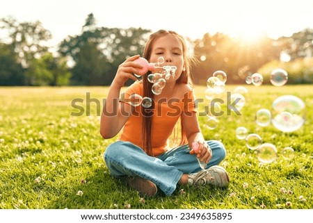 Cute beautiful girl blowing soap bubbles sitting on the grass in the meadow in the park, playing and having fun on a warm sunny weekend.
