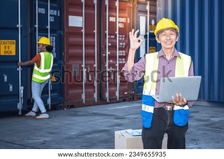 Asian man workers standing, holding a notebook is happy and use his hand to make an OK sign, with blur of African woman is opening containers background, to Container Cargo and Transportation concept.