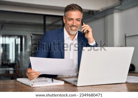 Smiling Hispanic businessman talking phone and check database in office. Happy Latin or Indian male business man holding documents, working at laptop computer doing online trade market tech research. Royalty-Free Stock Photo #2349680471