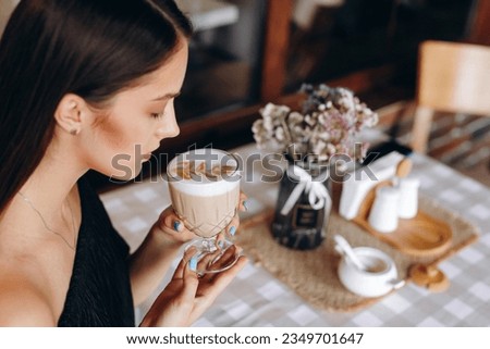Sophisticated woman holding a glass of Latte coffee in her hands and trying to take a sip on the background of a table with a checkered beige tablecloth, on which there is a brown cloth napkin