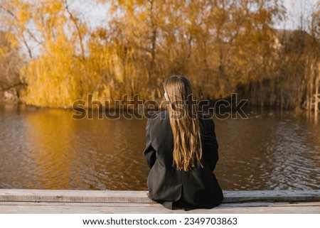 A woman with long hair in a black jacket is sitting on a wooden masonry near the lake in autumn. Back view. Royalty-Free Stock Photo #2349703863